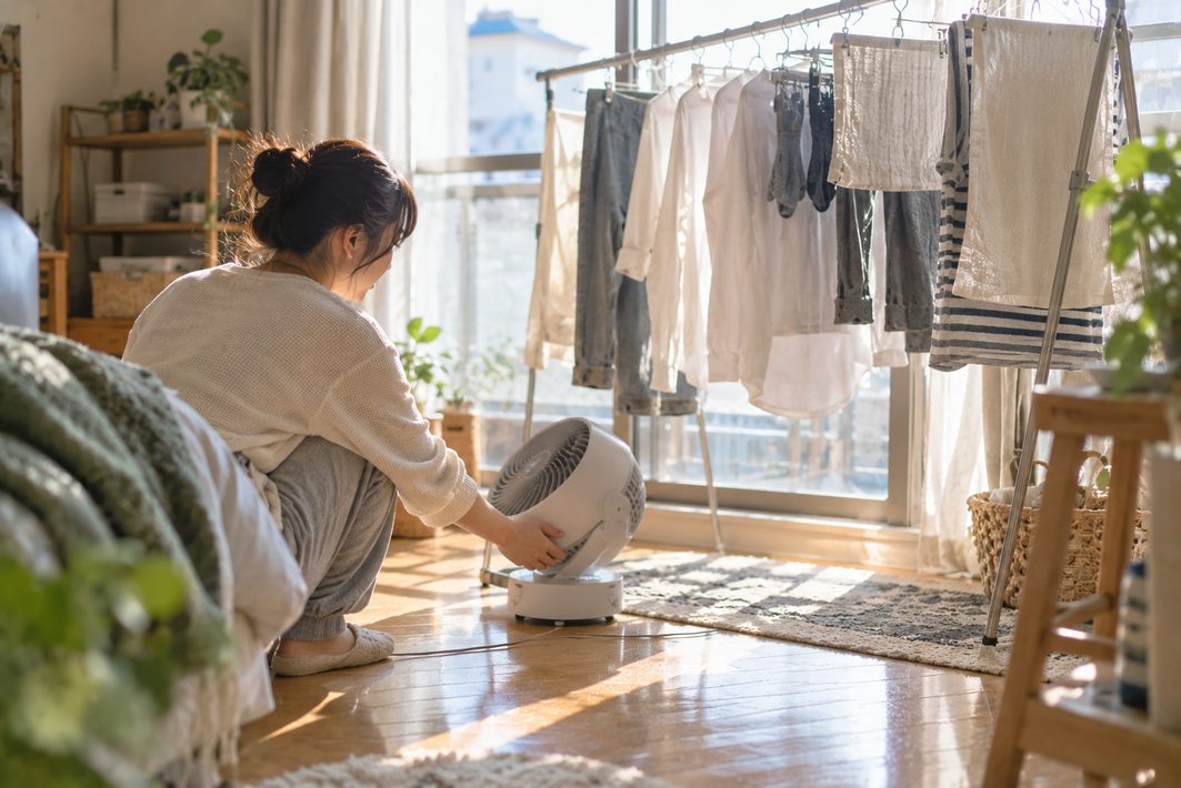 部屋干しの洗濯物のそばにサーキュレーターを置く女性